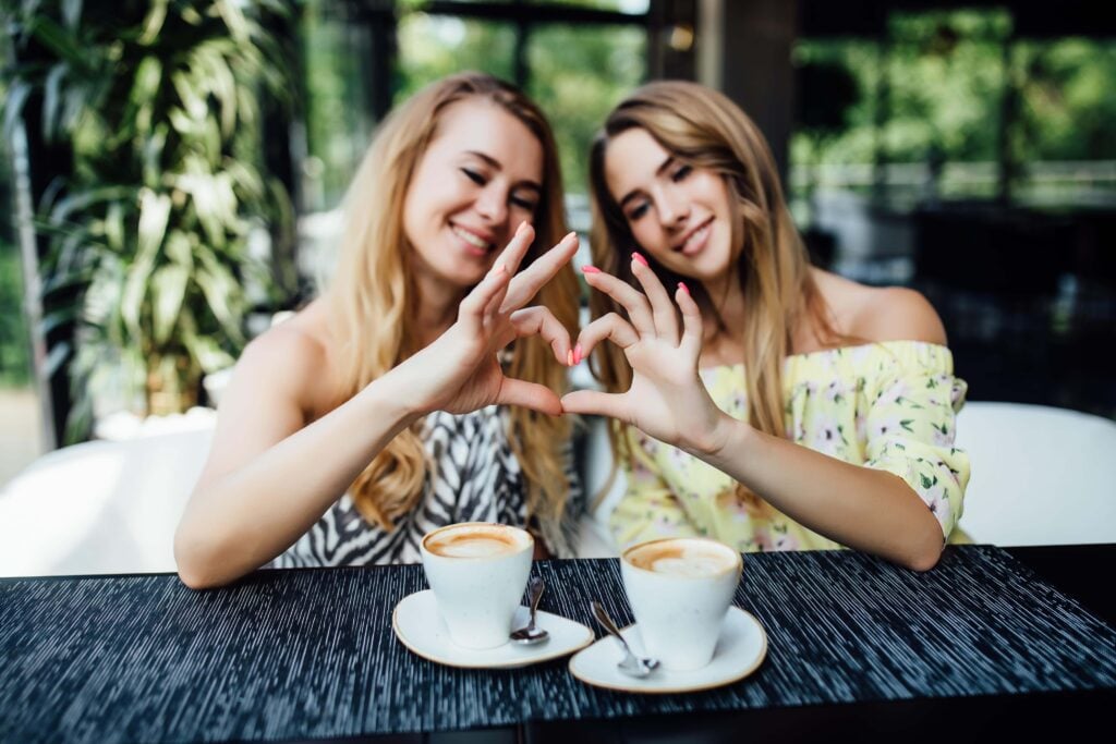 Two girl friends drinking coffee, speaking dutch phrases, and doing a heart shape with their hands